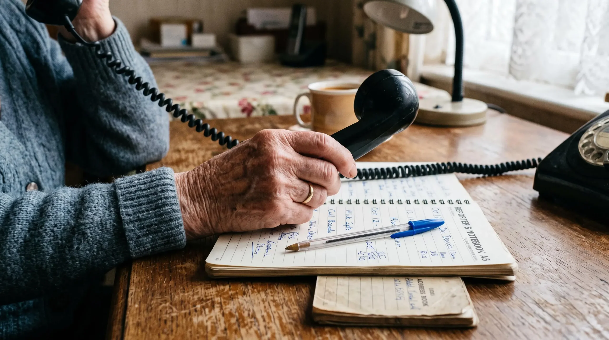 Gros plan sur une personne âgée passant un appel téléphonique, un carnet et un stylo posés sur la table.